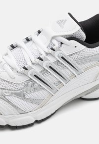 Balti sporta apavi ar sietu augšu, melniem akcentiem un sudraba detaļām. Iekļauj teksturētu zoli un izteiktu saistīšanas sistēmu.