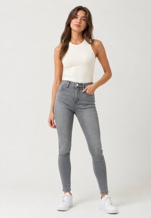 Frau in weißem ärmellosem Top, grauen hoch taillierten Skinny-Jeans und weißen Sneakern, stehend vor einem einfarbigen Hintergrund.