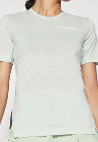 Vaaleanvihreä lyhytnenäinen urheilupaita, jossa on verkkosivupaneelit, pyöreä kaula-aukko ja valkoinen "TERREX"-logo yläetupuolella. Pehmeä kangastuntu.