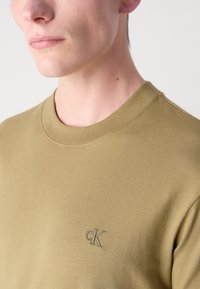 Olivově zelené bavlněné tričko s kulatým výstřihem a krátkými rukávy. Na horní levé straně se nachází decentní vyšívané logo "ck". Hladká textura.