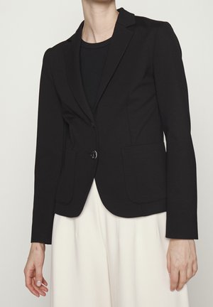 Person wearing black blazer over black shirt and white pants, standing against a plain light background.