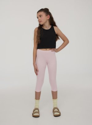 Ragazza giovane in piedi con una mano sui fianchi, indossa un top corto nero senza maniche, leggings capri rosa, calzini gialli e sandali beige.