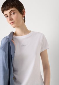 White short-sleeve t-shirt with a round neckline and rhinestone embellishment. A light blue blazer is draped over the arm.