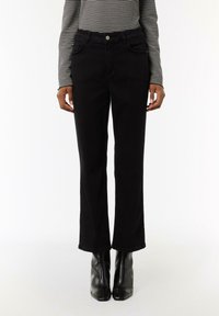 Schwarze, auslaufende Jeans aus Denim mit vorderen Taschen, einem Knopfverschluss und einer glatten Textur. Getragen mit schwarzen, hochhackigen Stiefeln.