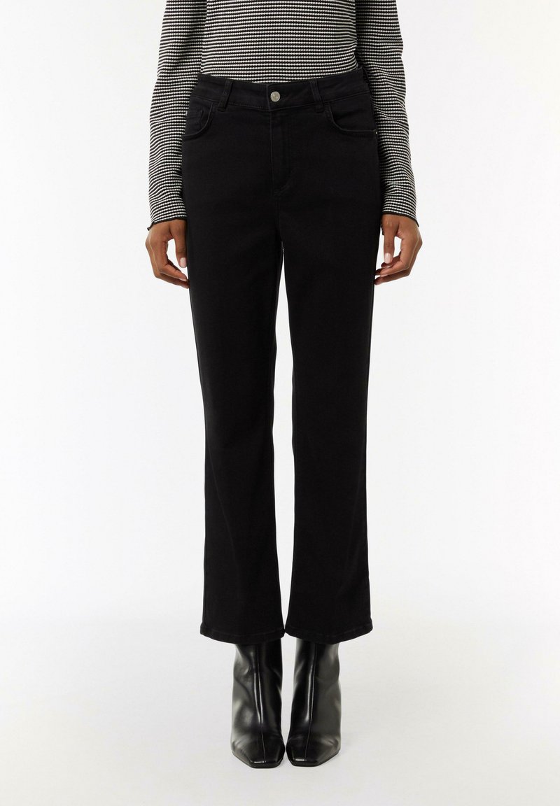 Schwarze, auslaufende Jeans aus Denim mit vorderen Taschen, einem Knopfverschluss und einer glatten Textur. Getragen mit schwarzen, hochhackigen Stiefeln.