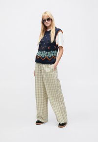 Gilet in maglia blu navy con motivo geometrico multicolore, abbinato a pantaloni ampi a quadri verde chiaro e scarpe nere. Occhiali da sole eleganti opzionali.