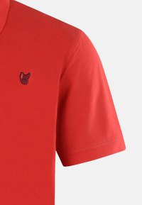 Röd polo t-shirt med korta ärmar, med en liten marinblå logotyp på vänster bröst. Slät textur och klassisk kragdesign.