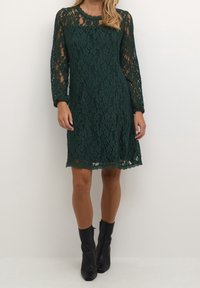Femme portant une robe en dentelle à manches longues vert foncé et des bottines noires, debout devant un fond blanc uni.