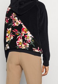 Czarna bluza z kapturem z kwiatowym materiałem po jednej stronie; posiada kieszeń kangurkę, ściągacz na dole i fakturowany materiał. Gładkie wykończenie w intensywnych kolorach.
