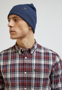 Granatowa, prążkowana czapka z małą czarną metką; noszona z wielobarwną koszulą w kratę, zawierającą odcienie czerwonego, niebieskiego i białego, z zapięciem na guziki.