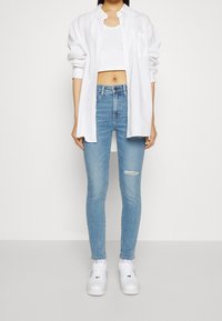 Camisa blanca de botones sobre un top corto blanco, combinada con unos jeans ajustados de mezclilla azul claro con rodilla desgastada y zapatillas blancas.