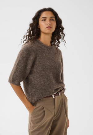Mujer con pelo rizado que lleva un suéter marrón de manga corta y peludo, metido en unos pantalones beige de talle alto con un cinturón marrón.