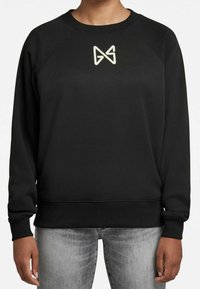 Persona con sudadera negra y logo geométrico claro en el pecho, usando jeans gris desgastado, de pie con los brazos relajados.