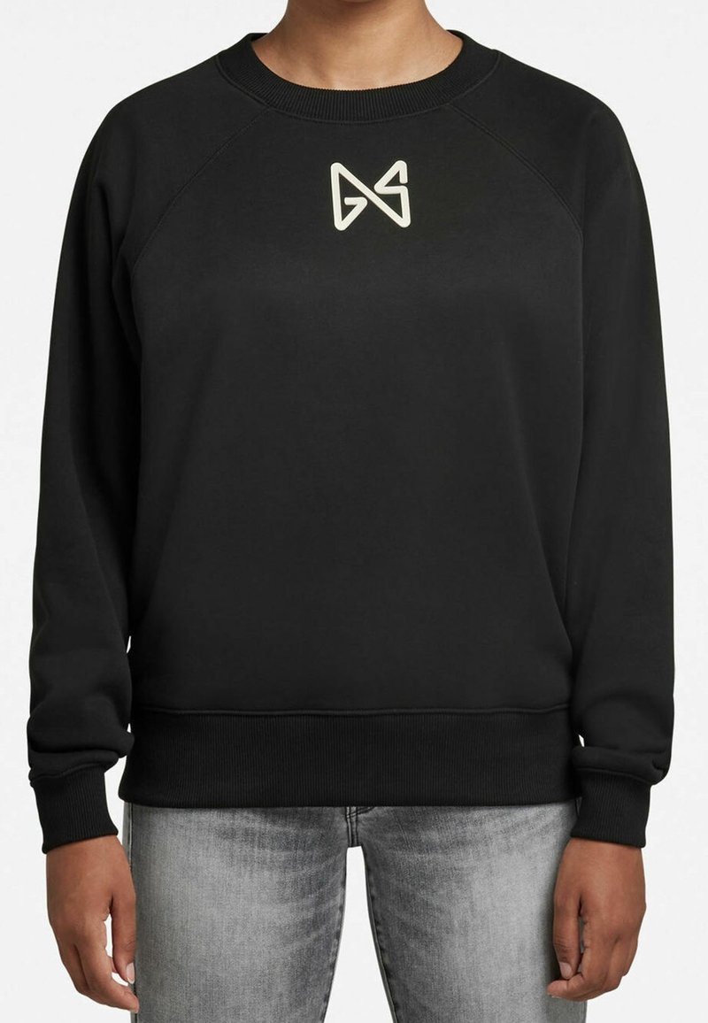 Persona con sudadera negra y logo geométrico claro en el pecho, usando jeans gris desgastado, de pie con los brazos relajados.