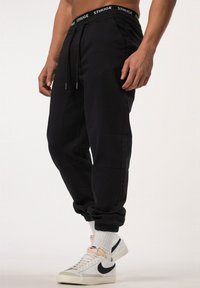 Pantalones de chándal negros con cintura elástica que presentan el texto blanco "STHUGE", cierre de cordón, puños ajustados y costuras laterales.