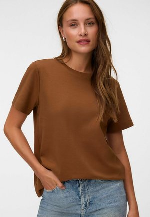 Kvinde med langt lysbrunt hår iført sølv hoops-øreringe, en brun kortærmet skjorte og lyseblå jeans, poserer med den ene hånd i lommen.