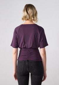 Lilla kortermet topp med avslappet passform og rynkete detaljer ved midjen, kombinert med svarte tettsittende jeans. Jevn stoffoverflate.