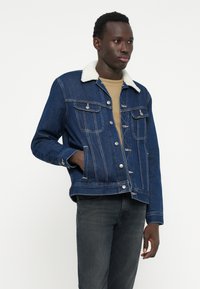 Jeans jakna v temno modri barvi z belim sherpa ovratnikom, z sprednjimi žepi, srebrnimi gumbi in sproščenim krojem. Nosite jo čez bež srajco.
