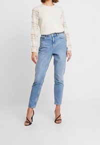 Krémový svetr s krajkovými dlouhými rukávy, v kombinaci se světle modrými džínami s vysokým pasem a černými sandály na vysokém podpatku. Hladké textury a přiléhavý design.