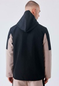 Chaqueta con capucha en negro y beige, que presenta un diseño a bloques de color, textura suave y mangas raglán. La vista trasera muestra un estilo liso y minimalista.