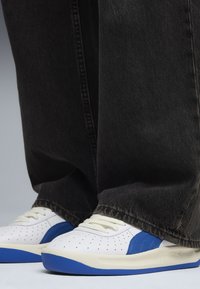 Weiße Sneakers mit blauen Akzenten, durchbrochenem Design und weißen Schnürsenkeln, kombiniert mit dunklen Jeans. Fokus auf die Details des Schuhwerks und den Farbkontrast.