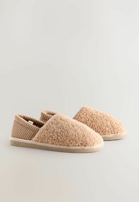 Beige Hausschuhe mit einer strukturierten, flauschigen Außenfläche und einem gerippten Stoffkragen. Weiche Sohle mit dezentem Branding an der Seite.