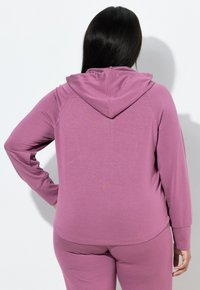 Felpa a maniche lunghe viola realizzata in tessuto morbido, con cappuccio e maniche raglan, abbinata a pantaloni coordinati per un completo.