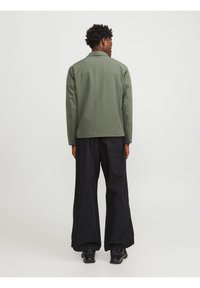 Veste verte à col, à manches longues, portée sur un pantalon noir ample, en tissu lisse, vue de dos.