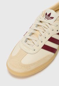 Cremefarvede lædersneakers med maroon striber, tekstureret suede tåhætte og hvide snørebånd. Har et tydeligt logo på tungen.