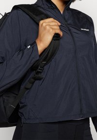 Marinblå lätt jacka med dragkedja framtill och hög krage, med en texturerad tyg och logodetalj på bröstet. Svart ryggsäckrem synlig.