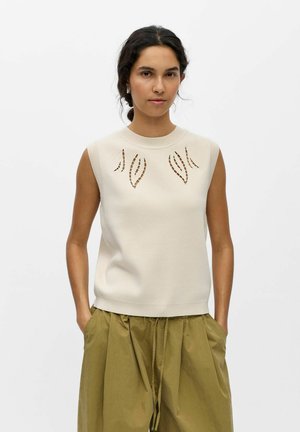 Frau trägt ärmellosen cremefarbenen Pullover mit ausgeschnittenen Blattmustern und lockere olivgrüne Hosen, steht mit Händen in den Taschen vor weißem Hintergrund.