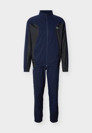 Navy blue and black full-zip track jacket with matching pants, elastic cuffs, and Lacoste crocodile logo on chest and thigh.