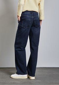 Jeans de denim azul oscuro con corte recto, que presentan dos bolsillos traseros y una textura desgastada. Combinados con zapatillas blancas.