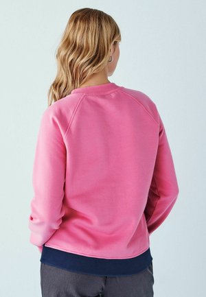 Femme aux longs cheveux blonds portant un sweat-shirt rose par-dessus une chemise bleu marine, debout de dos sur un fond clair uni.