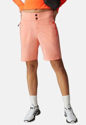 Persona que lleva pantalones cortos rosa claro con bolsillos laterales con cremallera, calcetines blancos hasta la media pierna y zapatillas blancas con detalles negros, de pie con las manos en la cadera.