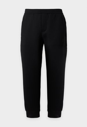 Νέος άνδρας με μπλε μπλουζάκι EA7 Emporio Armani, μαύρο παντελόνι και μαύρα αθλητικά παπούτσια, στέκεται μπροστά σε ένα απλό φωτεινό φόντο.