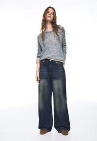 Dunkelblaue Jeans mit weitem Bein und verwaschenem Look, kombiniert mit einem hellgrauen Strickpullover. Der Pullover hat hochgekrempelte Ärmel und eine weiche Textur.