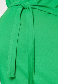 Zielona sukienka wykonana z gładkiego, lekkiego materiału z paskiem do związania. Jej fason charakteryzuje się dopasowaną sylwetką i widocznymi detalami przeszyć.
