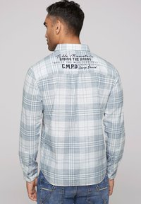 Blau-weiße Karobluse mit langen Ärmeln und abgerundetem Saum. Mit Text auf dem oberen Rücken und Zwei-Knopf-Manschetten.