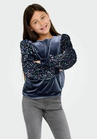 Sudadera azul de terciopelo con mangas abullonadas de lentejuelas, combinada con pantalones grises; presenta un escote redondo y un corte holgado.