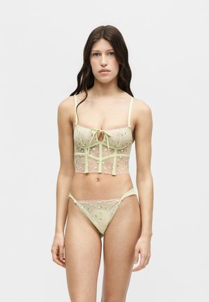 Young woman with long dark hair wearing pale green sheer embroidered lingerie set with a bustier top and matching bottoms standing against a plain background.