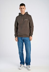 Mørkebrun hættetrøje med snore og frontlomme, parret med lyseblå jeans og hvide sneakers. Enkelt design, ingen yderligere mønstre.