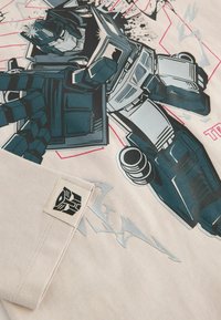 T-shirt in cotone beige con una grande grafica di un robot in nero e verde acqua, con effetti di spruzzo e un'etichetta con logo sull'orlo.