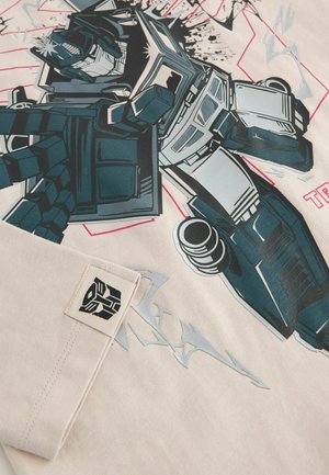 Camiseta de algodón beige con un gran gráfico de un robot en negro y verde azulado, con efectos de salpicaduras y un parche de logo en el dobladillo.