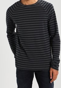 Chemise à manches longues gris foncé avec des rayures bleu marine. Tissu texturé, encolure ronde et manches raglan. Associée à un jean foncé.