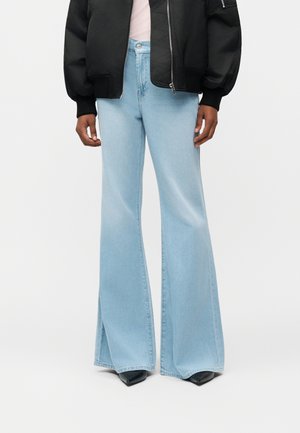 Osoba oblečená v svetlo modrých zvonových džínsoch, čiernych špicatých topánkach, ružovom topánku a čiernej bombera bunde so zipsovými vreckami.