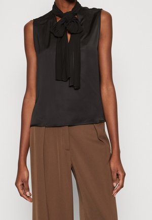 Mouwloze zwarte blouse met een gestrikte halsdetail, gecombineerd met hooggestemde bruine wijde broekspijpen gedragen door een staand persoon.