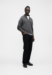 Gray zip-up sweatshirt with a high collar, worn over a white shirt, paired with black trousers and black shoes; features a small logo on the chest.