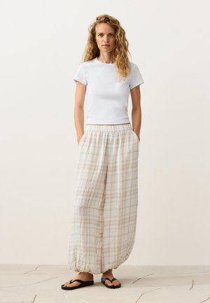 Žena s dlhými kučeravými vlasmi, ktorá nosí biele priliehavé tričko, voľné béžové kárované nohavice s elastickými manžetami a čierne žabky, stojaca na neutrálom pozadí.