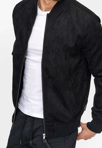 Veste bomber en daim noir avec une fermeture éclair à l'avant, des poignets côtelés et un design minimaliste. Portée sur un t-shirt blanc, avec une texture lisse.